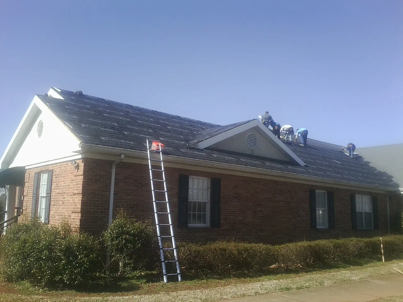 Crew tearing shingles off a brick ranch roof with a ladder set — Metal Roof Repair work by our crew in Hollis
