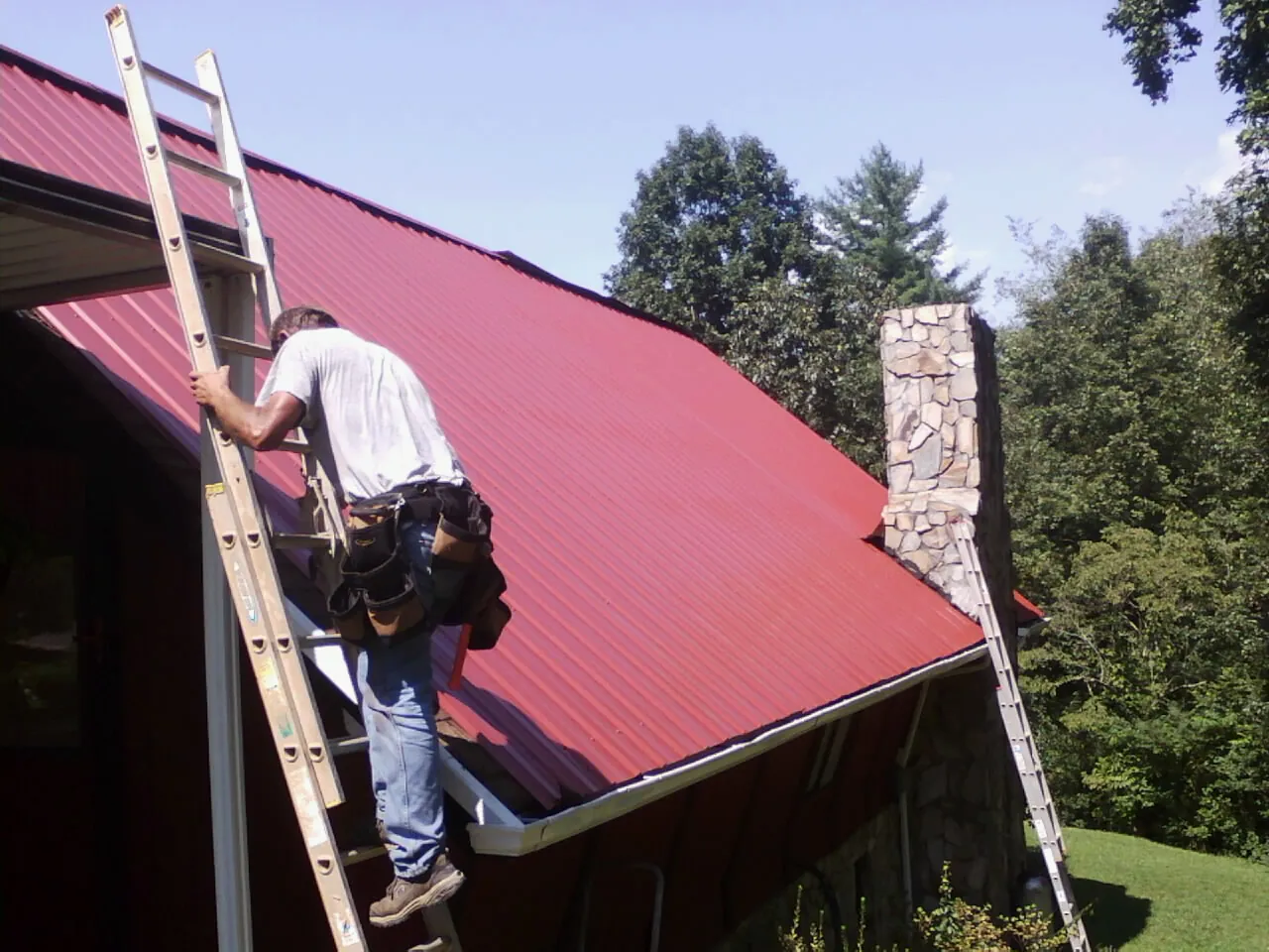 Roofer on a ladder beside a red metal roof and stone chimney — Soffit & Fascia Repair by Roofing Brothers in Hollis